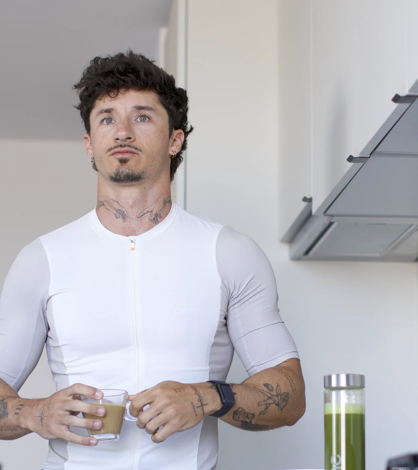 A cyclist in a bright kitchen holding a coffee beside a SEQUENCE green-juice bottle